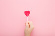 Little child hand fingers holding heart shape lollipop on stick on light pink table background. Pastel color. Sweet candy. Closeup. Top down view.