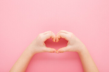 Wall Mural - Heart shape created from little girl hands on light pink table background. Pastel color. Love, health and hope concept. Closeup. Point of view shot. Top down view.