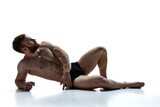 Fototapeta  - Full length portrait of young naked athlete man lying on floor against white background. Male model posing in underwear in studio. Concept of beauty care, male health, masculinity. Ad