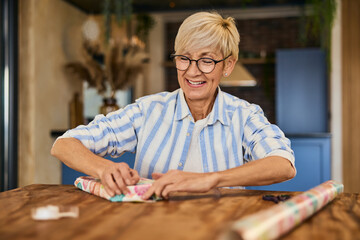 Wall Mural - A smiling mature woman, using decorative paper, wrapping a gift for someone, at home.