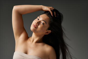 Wall Mural - young asian woman with blemishes and shiny brunette hair posing with hand in hair on grey background