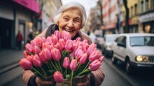 Portrait Of A Happy Mature Woman Standing On The Street With A Bouquet Of Tulips. Grandma Sells Tulips For The Holiday. Women's Day, Valentine's Day.