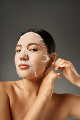 Wall Mural - attractive young asian woman applying hydrating sheet mask for skin treatment on grey background