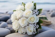  A Bridal Bouquet Of White Roses Sits On A Rock On The Beach With The Ocean In The Background Of The Photo And A Book In The Foreground.