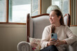 Mujer mirando por la ventana sonriente mientas usa su celular, sentada cómodamente en su sillón