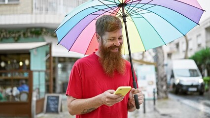 Wall Mural - Confident young redhead man joyfully smiling, holding an umbrella and using a smartphone on a bustling city street.