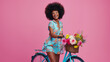Portrait of a beautiful African American Black woman with an Afro on a bicycle with a flower basket over pink studio background. Health, active, fitness, wellness, summer, spring, flowers, bloom
