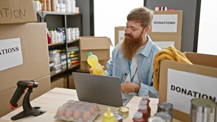 Sticker - Hardworking young redhead volunteer man holding a box of food donations, joyfully chatting on a video call indoors at the bustling charity center