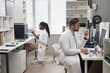 Side view of Caucasian male technician holding 3D dental model and looking at computer screen while sitting at desk in laboratory, Black female colleague working in blurred background