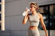 Young blonde athlete girl drinking water from sports bottle during morning jogging in the city. Female fitness model monitors the body water balance to avoid dehydration. Healthy lifestyle.