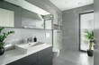 The image shows a modern bathroom with grey tiles, a white sink, mirror, toilet, and a glass shower. There are two green plants for a natural touch. Natural light comes in through a window.