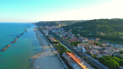 Wall Mural - Relaxed aerial push-in shot from Cupra Marittima (Ascoli Piceno) with undulating Adriatic Sea, breakwaters, sandy beach, resorts, parks, railway, buildings, and a natural Marche hillscape with trees