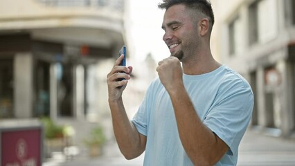 Wall Mural - A young hispanic man with a beard smiles while using a smartphone on a sunny urban street.