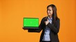 © DC Studio - Business person holds laptop with greenscreen on camera, showing blank copyspace display on portable pc. Asian woman in formal suit presents computer with chromakey screen in studio.