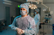 © dikushin - Portrait of confident male doctor in surgical mask and hat in operating room. Practitioner standing posing looking at camera in hospital, team of diverse surgeons performing operation on background