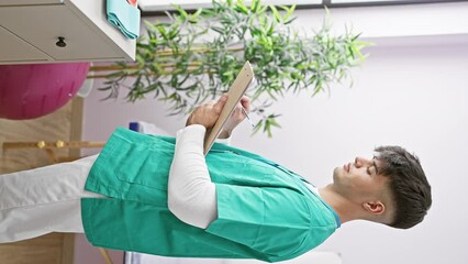 Canvas Print - Handsome young hispanic man, a smiling physiotherapist, diligently taking notes at a bustling rehab clinic