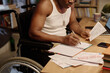 © AnnaStills - Unrecognizable black man in wheelchair writing information from piece of paper into his notebook