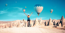 Woman Tourist In Bicycle Enjoying Cappadocia Landscape With Hot Air Balloons