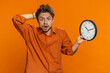 Caucasian man with anxiety checking time on clock, running late to work, being in delay, deadline. Young guy looking at hour, minutes, worrying to be punctual isolated on studio orange background