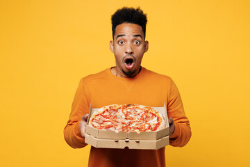 Wall Mural - Young surprised man wear orange sweatshirt casual clothes holding Italian pizza in cardboard flatbox isolated on plain yellow background. Proper nutrition healthy fast food unhealthy choice concept.