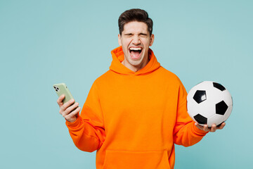 Young man fan wears orange hoody casual clothes use mobile cell phone scream cheer up support football sport team hold in hand soccer ball watch tv live stream isolated on plain blue color background.