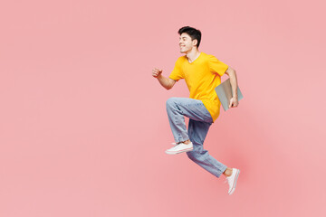 Wall Mural - Full body young IT man he wear yellow t-shirt casual clothes jump high hold closed laptop pc computer run fast isolated on plain pastel light pink color background studio portrait. Lifestyle concept.