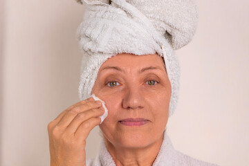 Wall Mural - beautiful elder old woman granny is applying face cream,use cotton disk and hydrofil oil to remove makeup.female grandmother looking in double sided mirror against wall, dressed in bath robe 4k