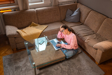 Top view of young pensive Caucasian woman wearing glasses is sitting near sofa and using laptop. Concept of remote work and freelance