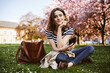 © contrastwerkstatt - Young Student Sitting with Smartphone in Spring Park, Leaning Head on Hand, Looking into Camera
