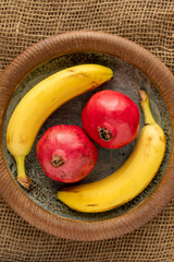 Yellow and red fruits: two pomegranates and bananas on a handmade clay plate. Jute fabric background. Top view.