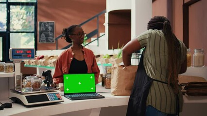 Vendor using a laptop with greenscreen layout at store checkout, serving regular customer with natural products. Women chatting next to pc showing isolated display and mockup copyspace. Tripod shot.