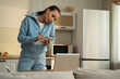 © Grustock - A young woman is standing in the kitchen , writing in a notebook using a laptop.