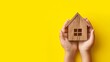  The childs hands hold a wooden house on a yellow background. The concept of adopting orphans into families, as well as purchasing and lending housing.