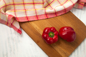 Cutting board with two whole red bell pepper and red kitchen towel on white wooden background. .