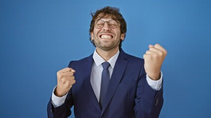 Wall Mural - Joyful young man in business suit, hispanic and blond, celebrates triumph arms raised. epic win, beaming with success! isolated on a blue background.