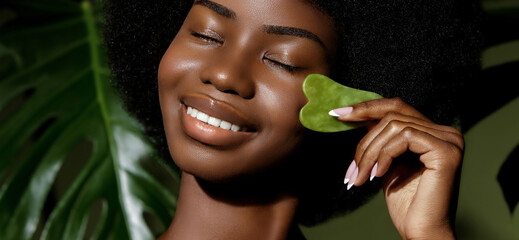 Wall Mural - gua sha jade skin treatment. african american model making face massage with a stone tool against tr