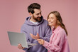 Couple of smiling friends holding laptop computer choosing goods isolated on pink background. Stylish man and woman shopping online looking at screen. Technology concept