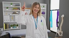 Joyful Middle Age Blonde Physio Therapist Strikes A Thinking Pose In Rehab Clinic, Her Confident Face Lighting Up With A Smart Idea, As She Pointedly Raises One Finger With A Cheeky Smile!