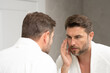 a man in a white bathrobe in the bathroom looking at the skin on his faceSpa male model, skincare product for skin face. Skin care. Male face cream. Man with perfect soft skin. Beauty cosmetic product