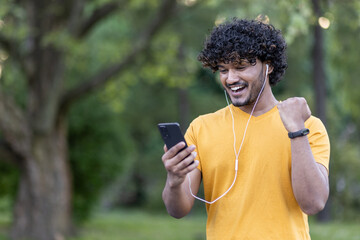 Indian young man doing sports in the park, standing in headphones and happy looking at phone screen showing victory hand gesture yes