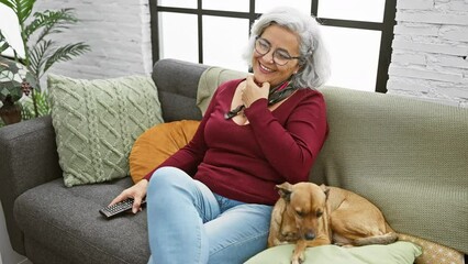 Poster - smiling woman with glasses and grey hair relaxes on a sofa with her pet dog in a cozy living room.