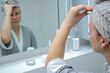 © TatjanaMeininger - Young woman in bathrobe and towel looking in a mirror in bathroom, making facial massage with a guasha