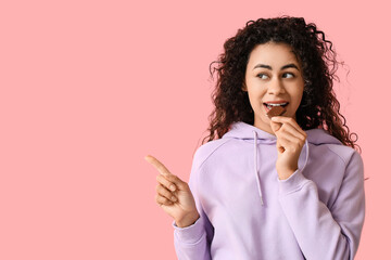 Canvas Print - Beautiful young African-American woman eating piece of sweet chocolate and pointing at something on pink background