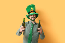 Young Man In Leprechaun Hat With Green Beard Holding Bottles Of Beer On Yellow Background. St. Patrick's Day Celebration