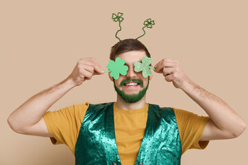 Wall Mural - Young man in headband with green beard holding clover on beige background. St. Patrick's Day celebration