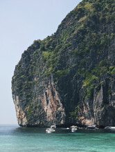 A Massive Rocky Cliff Surrounded By Turquoise Waters. The Cliff Is Covered With Green Vegetation And Has Rugged Textures. Several Boats Are Anchored Near The Base Of The Cliff. 