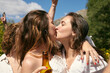 © Just images - A tender moment of a friendly kiss on lips between two women amidst the joy of a sunny outdoor gathering, with one holding a beer bottle.