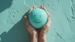 Overhead view of two hands carefully holding a smiley-faced stress ball, offering a sense of joy and relief against a textured turquoise backdrop.
