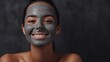 Spa facial treatments for woman. Dark skinned brunette happy girl smiles with a clay face mask on a dark gray background.  Take care of your skin. The health, beauty and youth concept