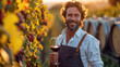 © Katerina Bond - A middle-aged man winemaker holds a glass of red wine and smiles, against the backdrop of a vineyard and barrels of wine. Winery, spirits production, sommelier.